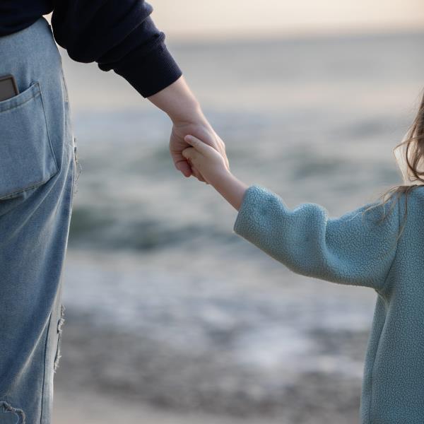 En voksen og et barn holder hender. Hendene er i fokus, de er vendt mot sjøen og stranda du ser i bakgrunnen.