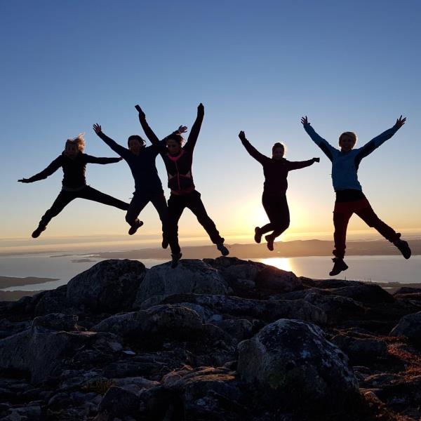 Fem damer hopper i lufta må toppen av Høvikfjellet med solnedgangen i bakgrunnen.