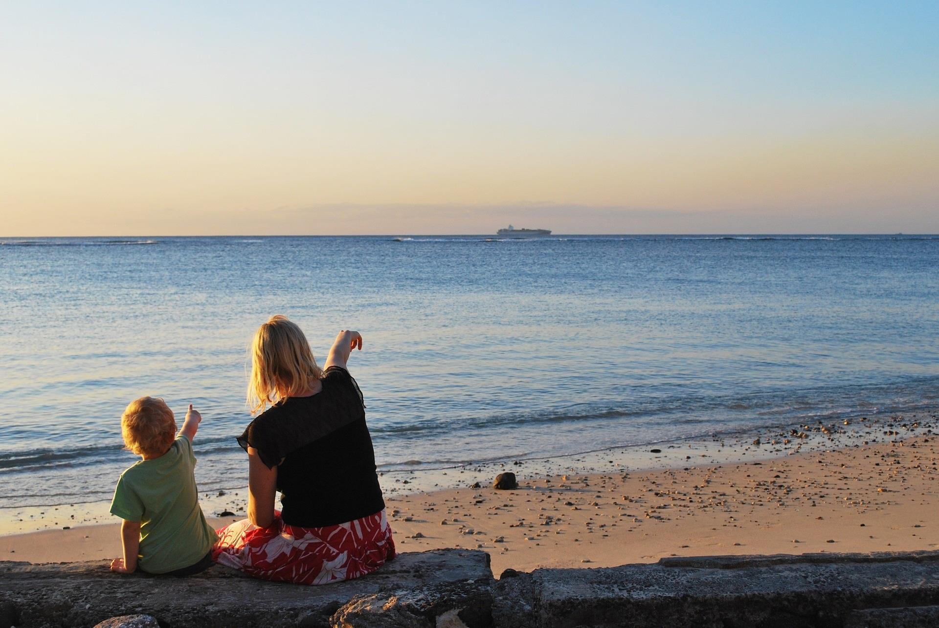 En mor og et barn sitter på stranda og peker utover havet. - Klikk for stort bilde
